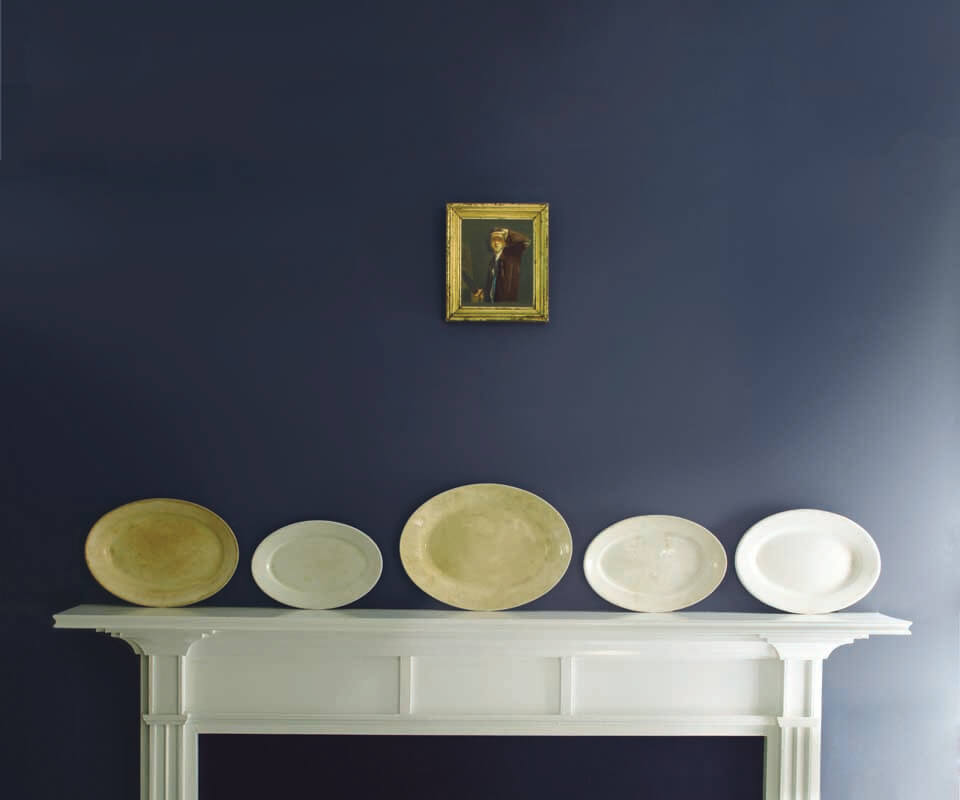 Plates on a white mantelpiece beneath a small portrait on a blue wall.